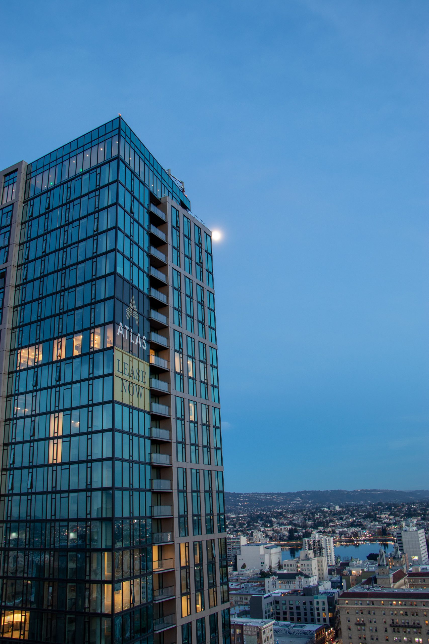 Modern glass skyscraper at dusk overlooking a sprawling cityscape with a visible moon nearby.
